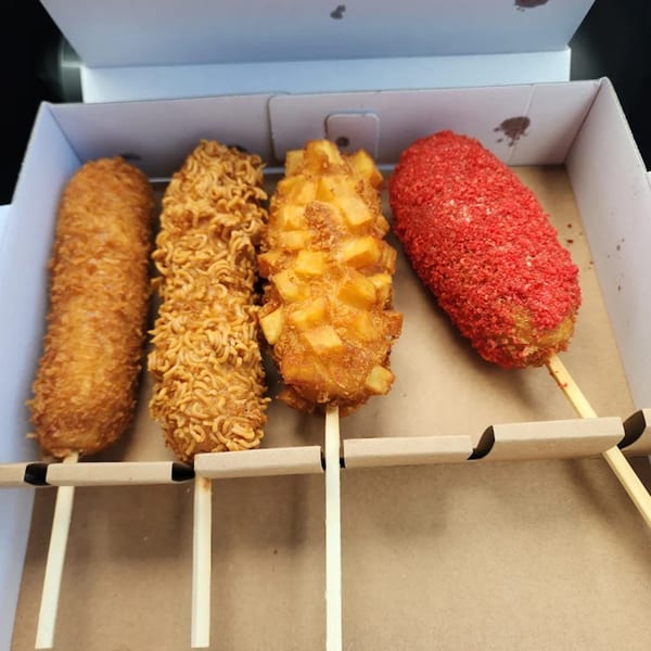 Four Daddy Donut corn dogs with various toppings, including ramen and red spice, in a white box.