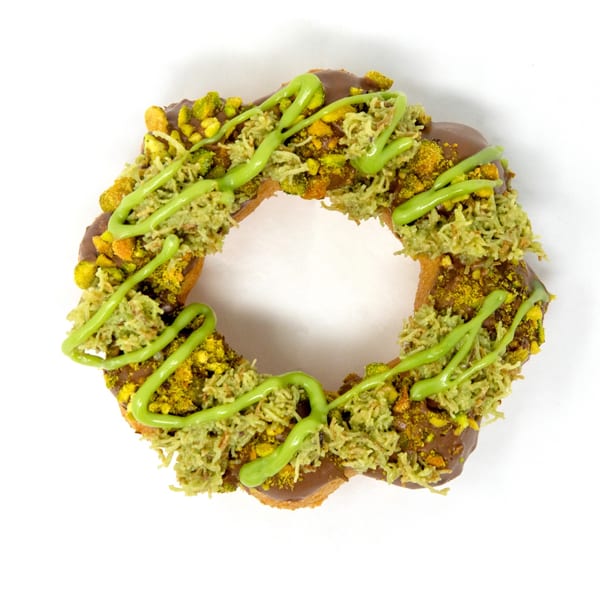 A Daddy Donut with chocolate frosting, green pistachio crumbles, shredded coconut, and green icing drizzle.